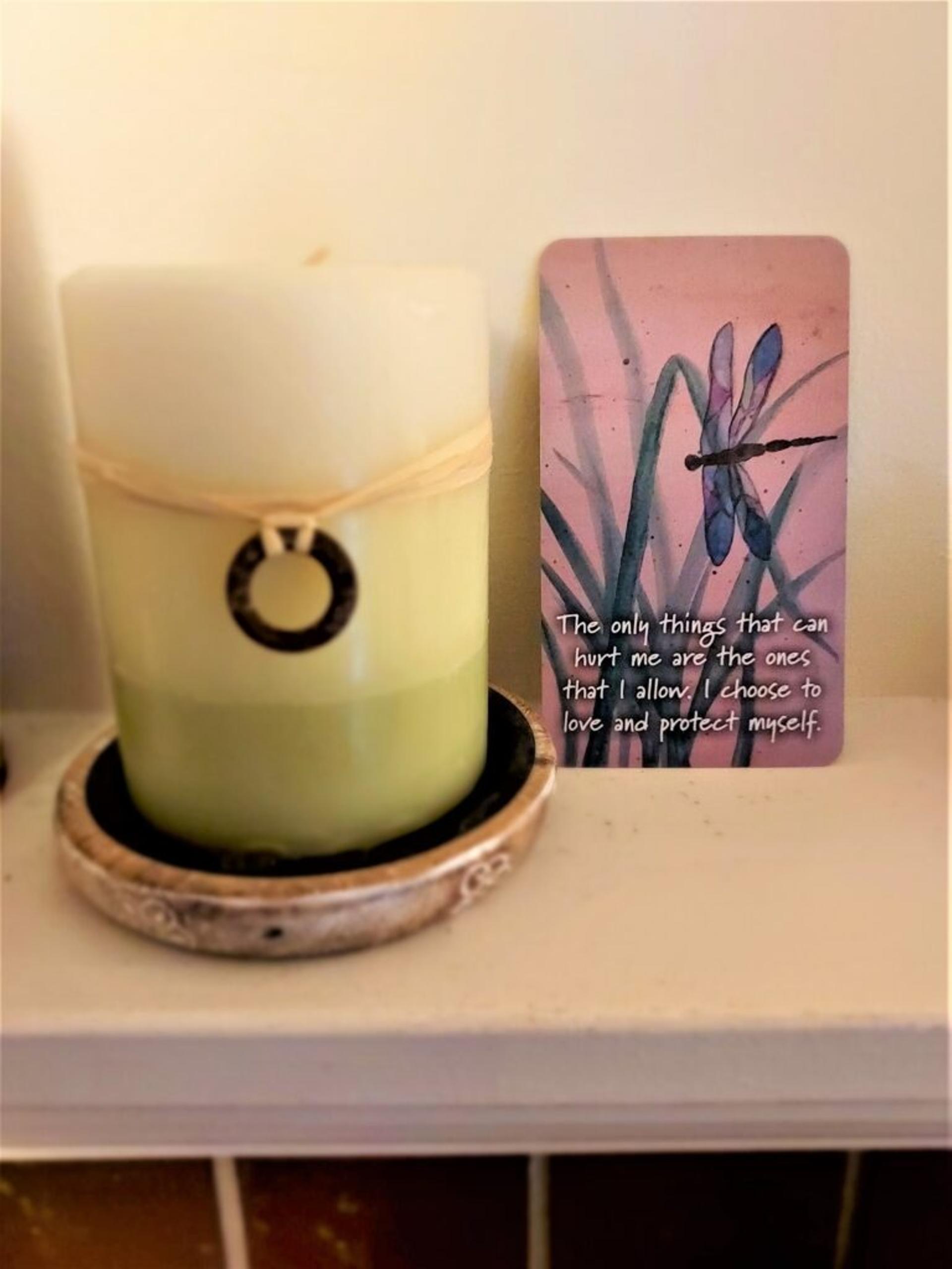 Card rests next to a candle; shows a watercolor painting of a dragonfly with text: "The only things that can hurt me are the ones that I allow. I choose to love and protect myself."