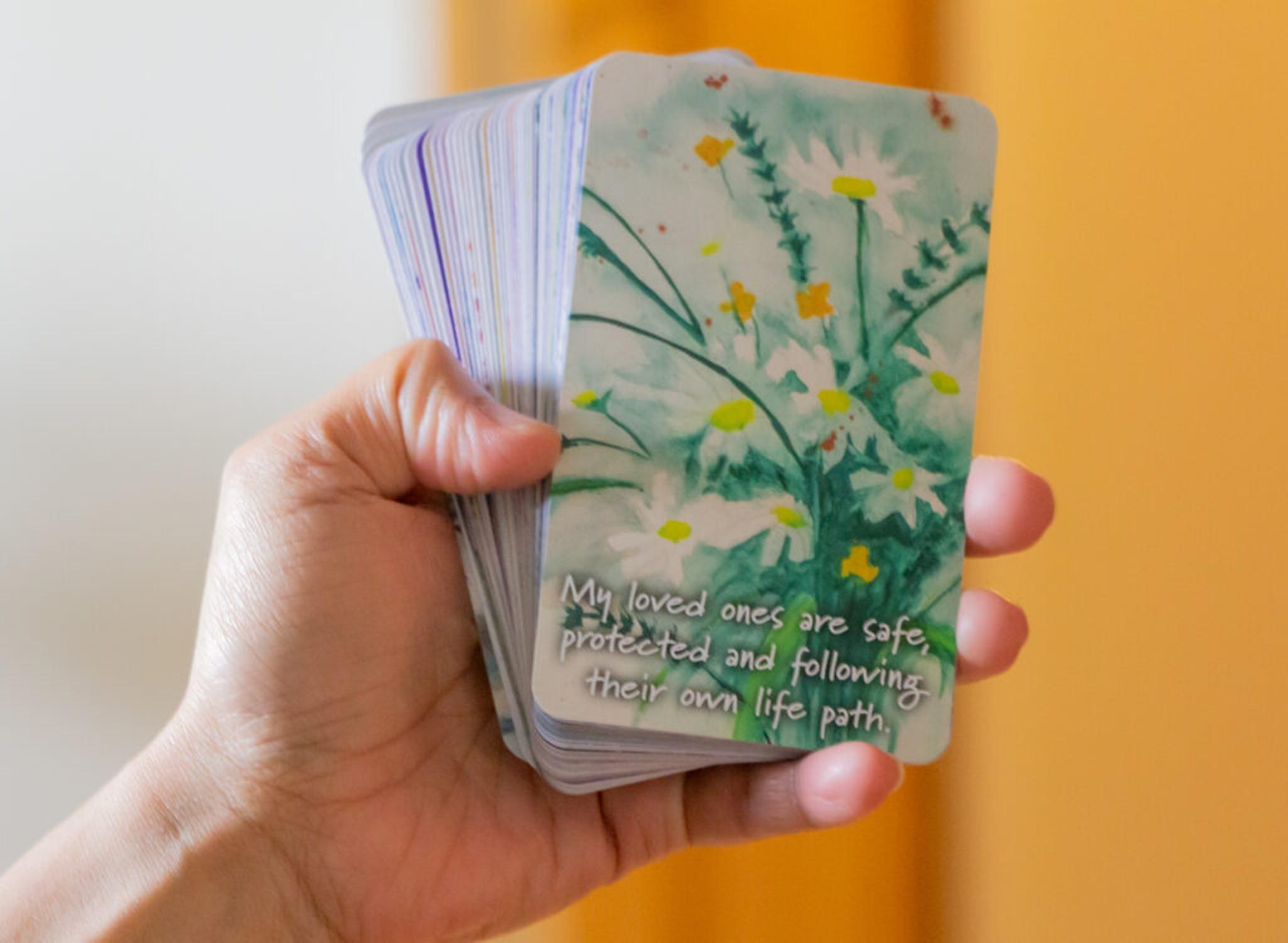 Hand holding deck of cards. First card shows a watercolor painting of daisies with text: "My loved ones are safe, protected and following their own life path."