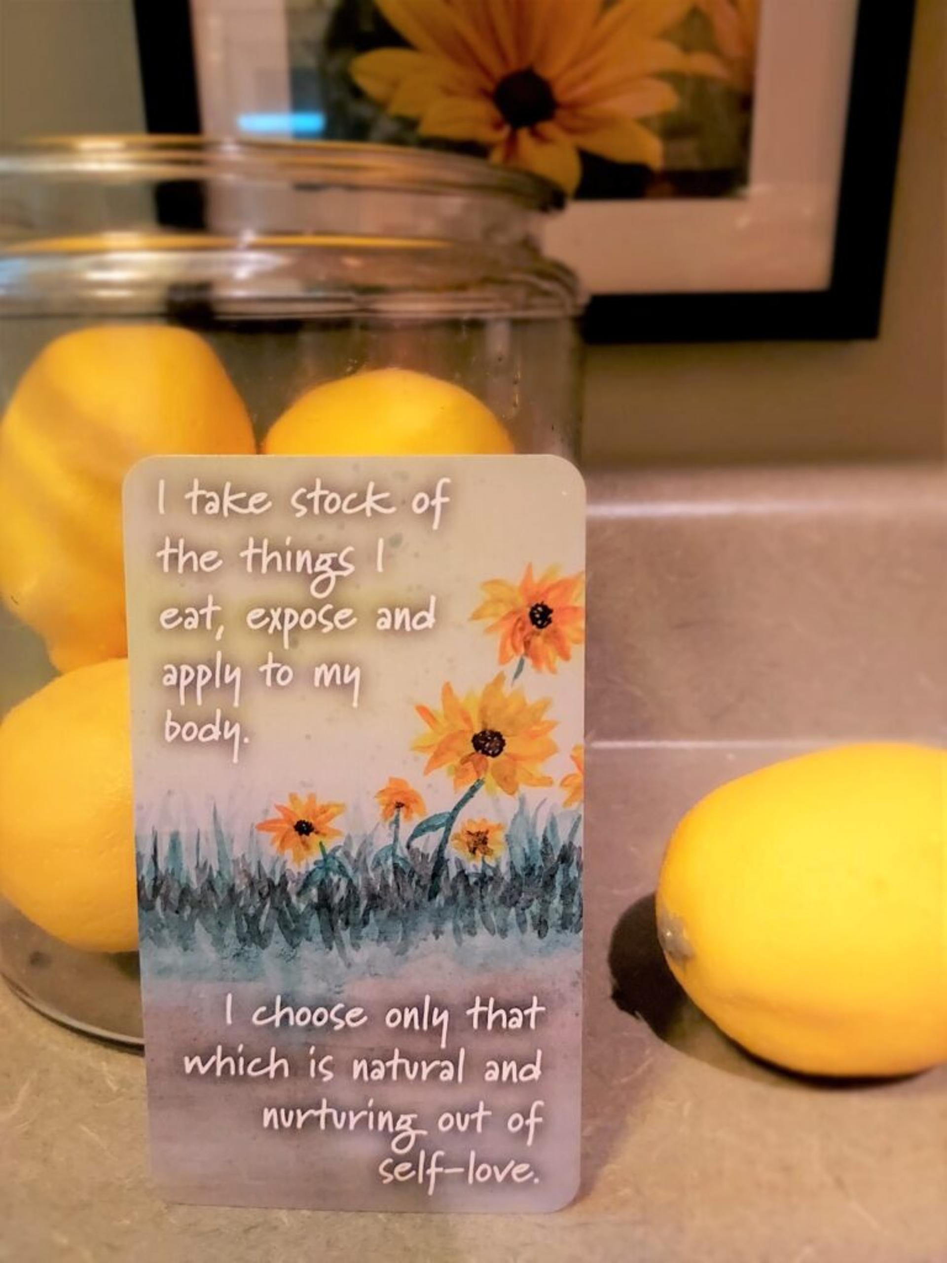 Card rests next to a jar of lemons; shows a watercolor painting of brown-eyed susan flowers with text: "I take stock of the things I eat, expose and apply to my body. I choose only that which is natural and nurturing out of self-love."