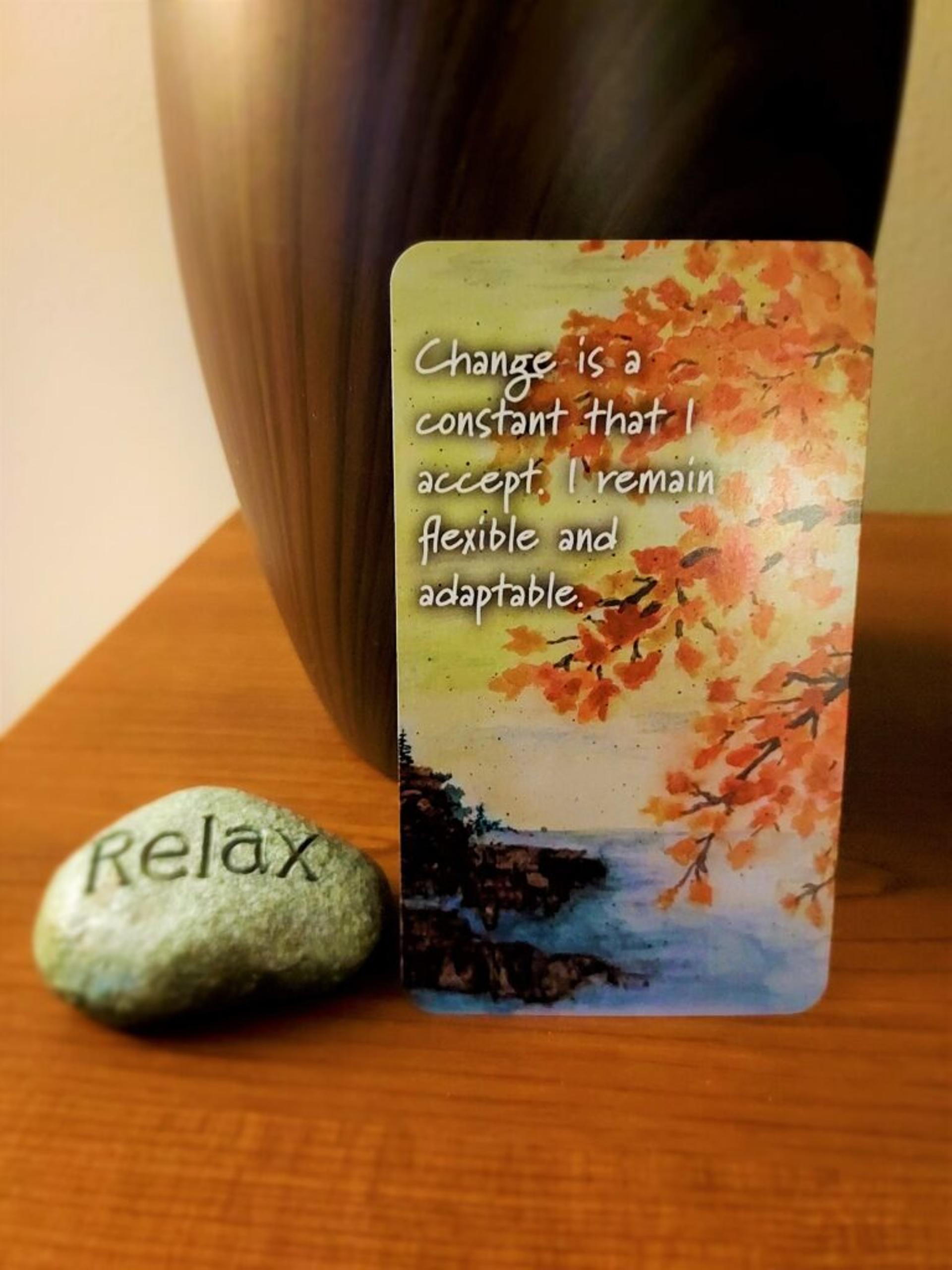 Card rests next to a stone engraved with "relax"; shows a watercolor painting of an autumn tree next to a shoreline with text: "Change is a constant that I accept. I remain flexible and adaptable."