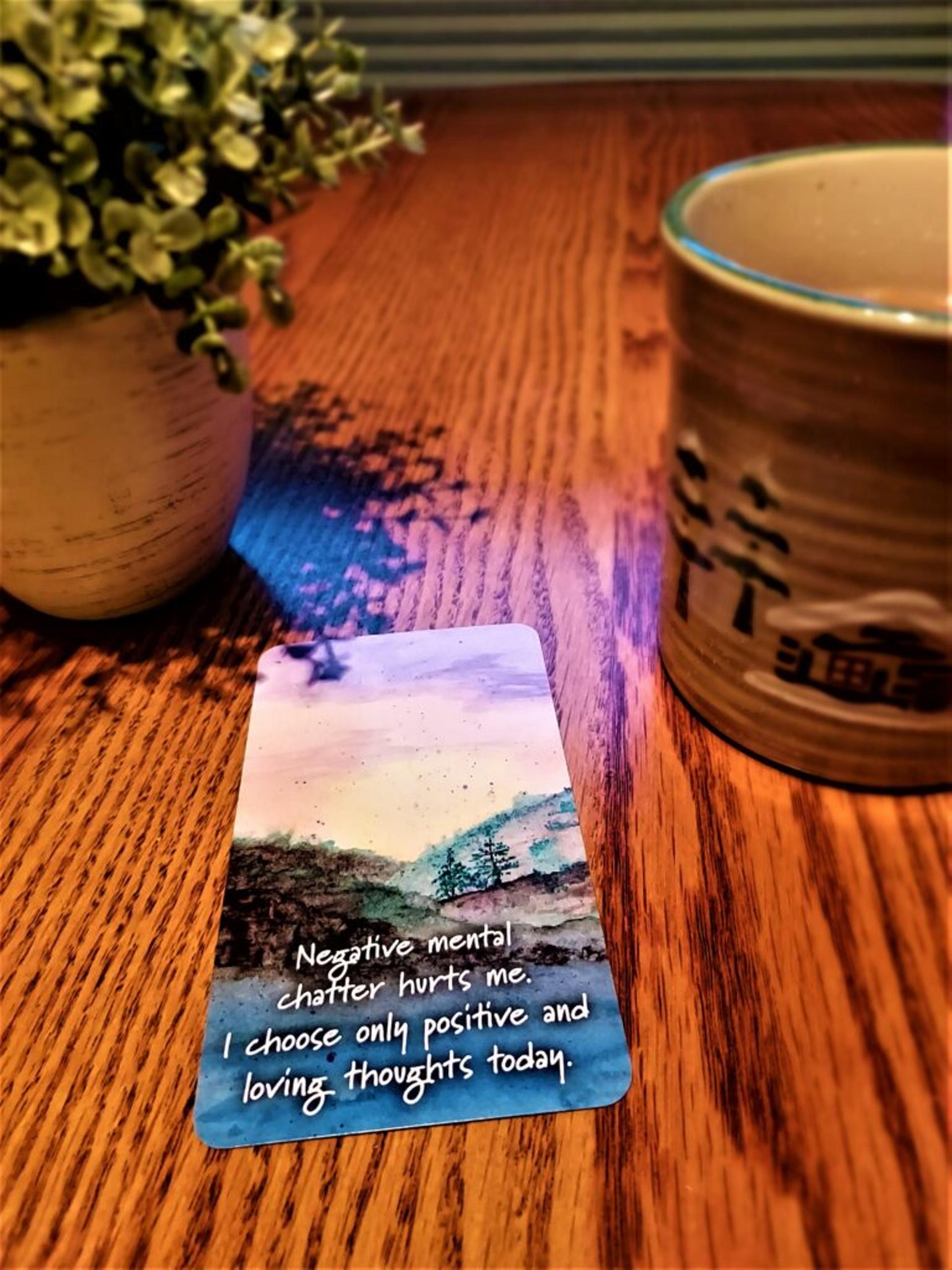 Card rests next to a mug; shows a watercolor painting of a landscape with text: "Negative mental chatter hurts me. I choose only positive and loving thoughts today."